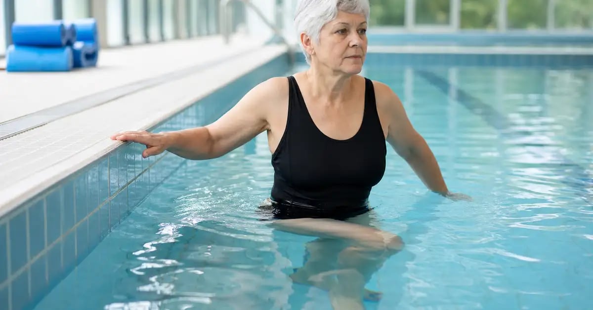 An older patient performing knee-bending exercises in a swimming pool during an aquatherapy rehabilitation session for knee osteoarthritis